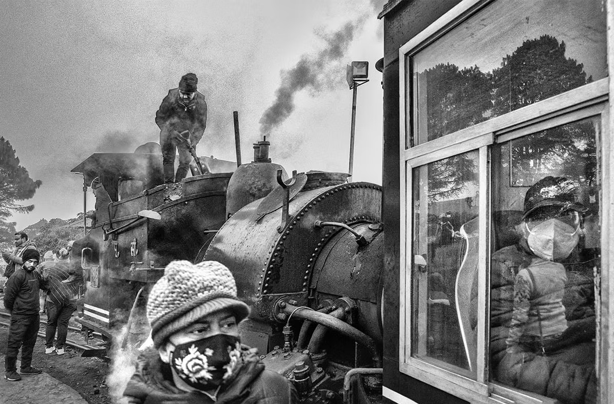 Arpan Dutta Chowdhury: Black, White & In Between 4 Workers tend a steam locomotive as masked passengers look from a carriage window, black-and-white street scene.