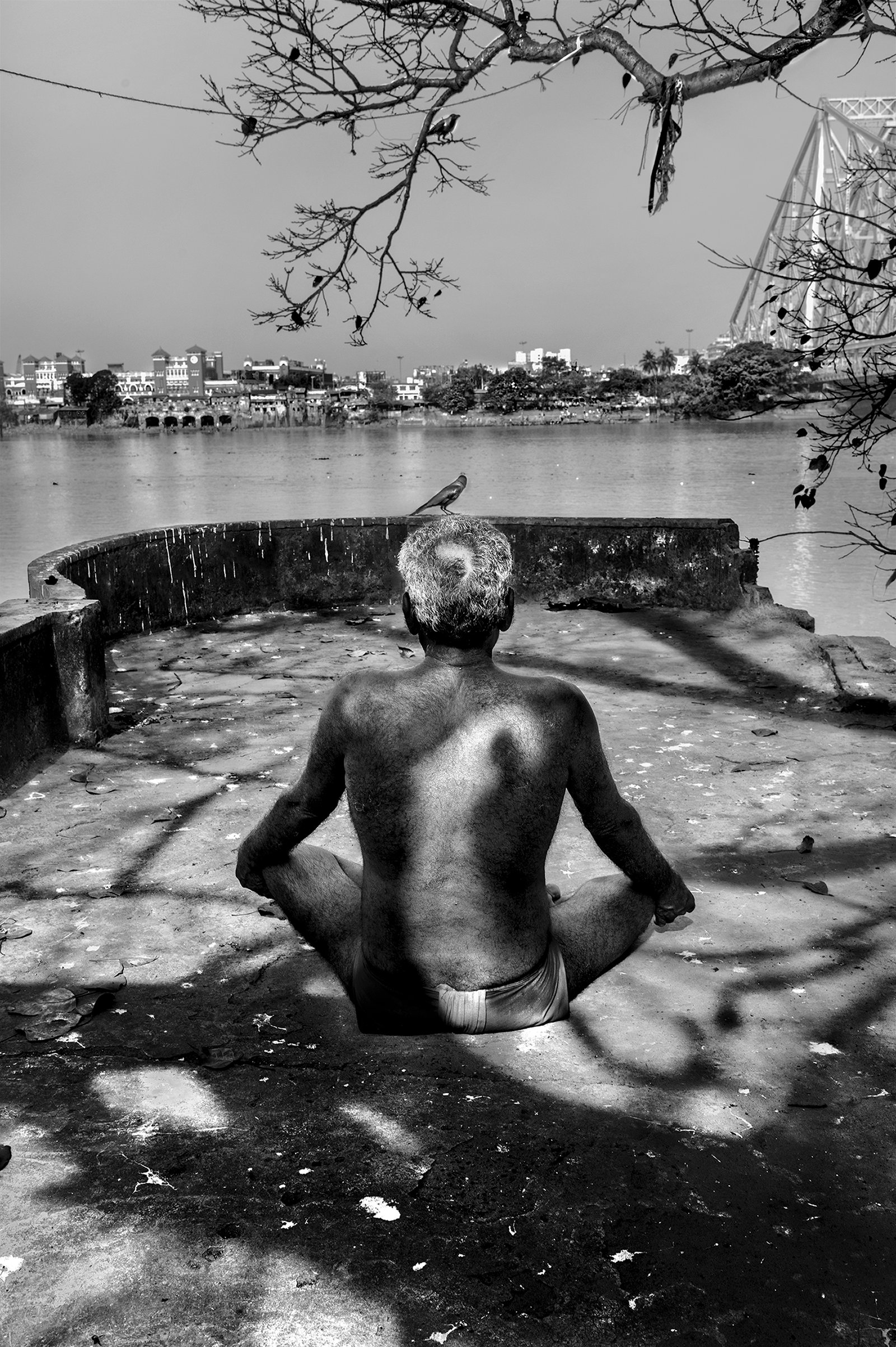 Arpan Dutta Chowdhury: Black, White & In Between 1 Elderly man sits bare-backed at a riverside terrace beneath branches, distant bridge and city across the water, B&W.